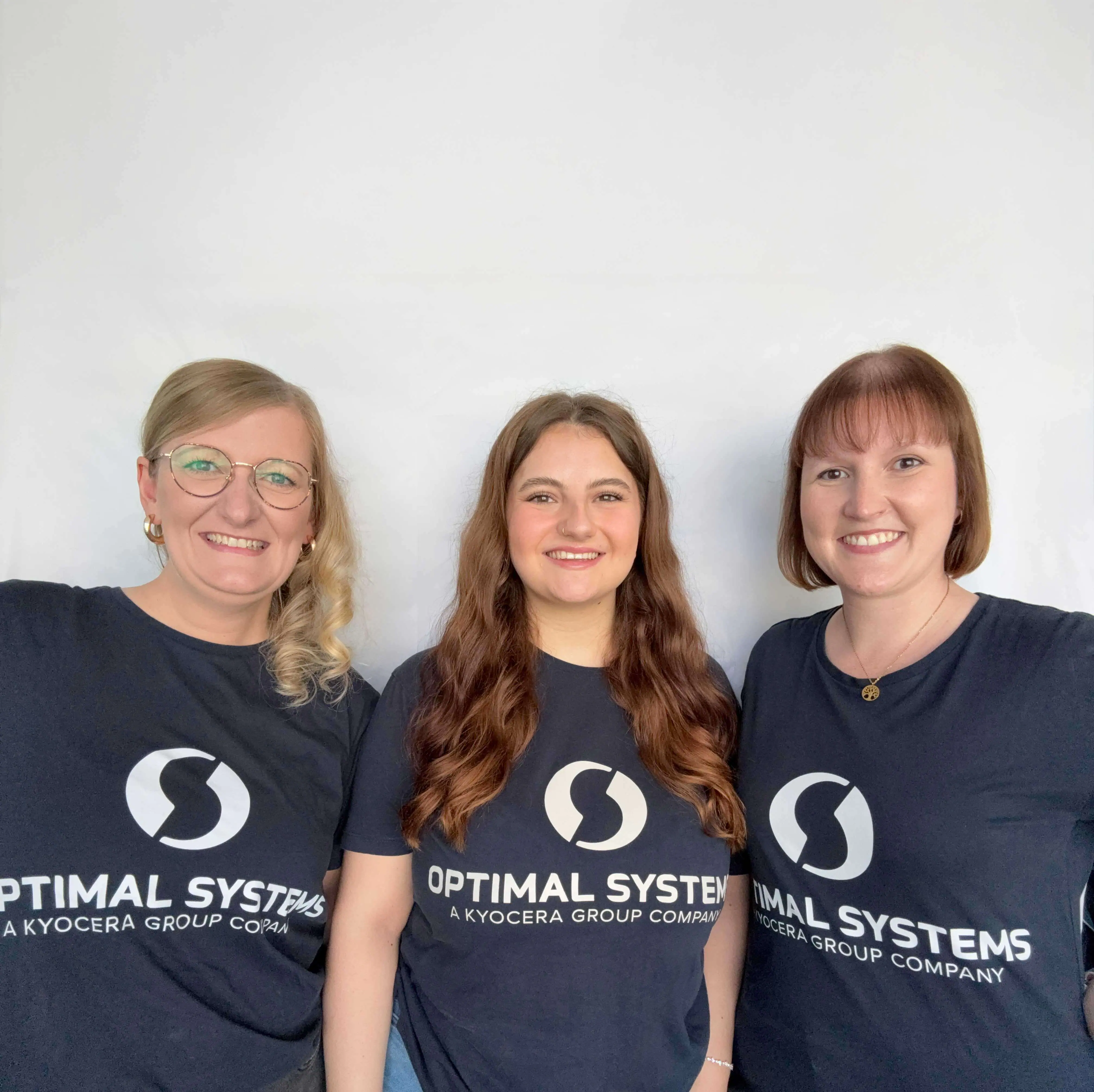Drei Frauen lächeln in die Kamera. Sie tragen passende marineblaue "OPTIMAL SYSTEMS"-T-Shirts, das weiße Logo ist sichtbar, vor einem schlichten hellen Hintergrund.