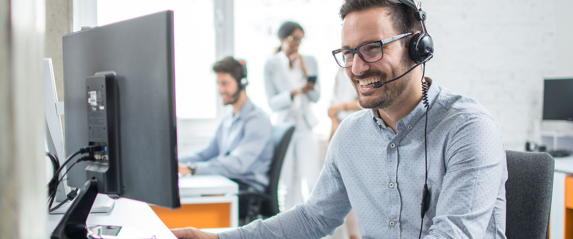 Lachender Mann mit Brille und Headset vor einem Computer mit Kollegen im Hintergrund