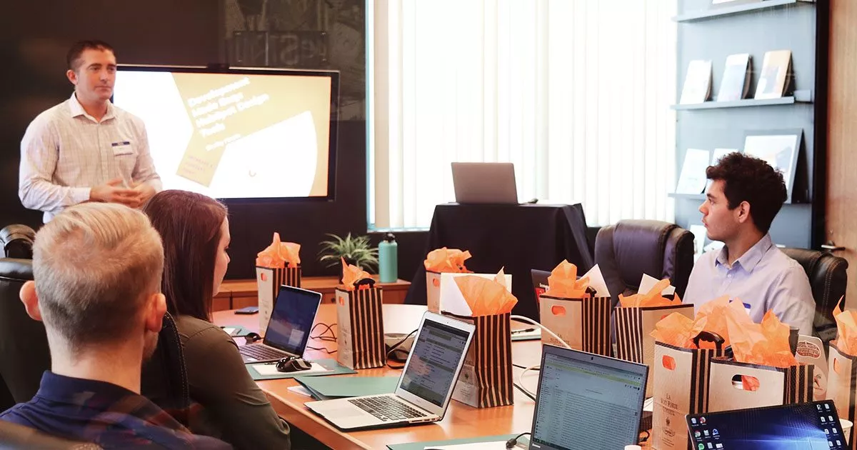 Eine Gruppe von Menschen sitzt mit Laptops an einem Tisch, während ein Mann einen Vortrag hält