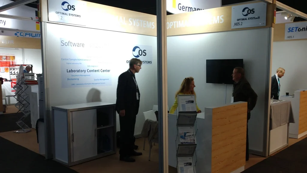 Exhibition booth for Optimal Systems featuring two people in conversation with branding and product information displayed on the booth wall.