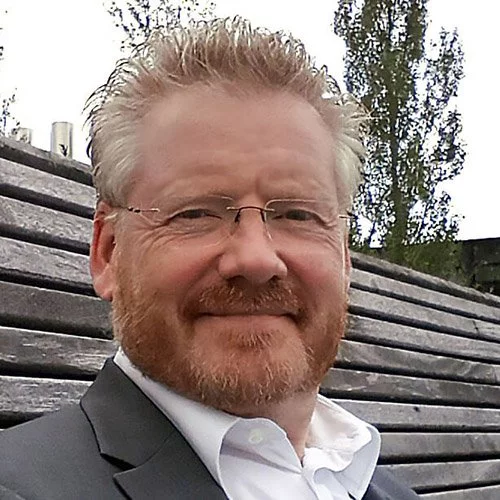 Ein Mann in Anzug und Krawatte steht lächelnd vor einem Holzzaun und präsentiert mdr insellösungen.