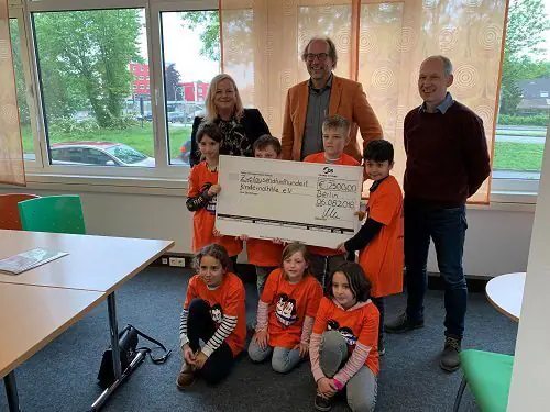 Group of adults and children holding a large check for a donation, with children wearing orange shirts and smiling for the photo.