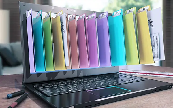Auf einem Schreibtisch steht ein geöffneter Laptop. Bunte, digitale Ordner scheinen aus dem Bildschirm herauszuspringen. Auf dem Schreibtisch liegen Stifte, im Hintergrund sieht man unscharfe Möbel. Das Bild symbolisiert die digitale Dateiorganisation.