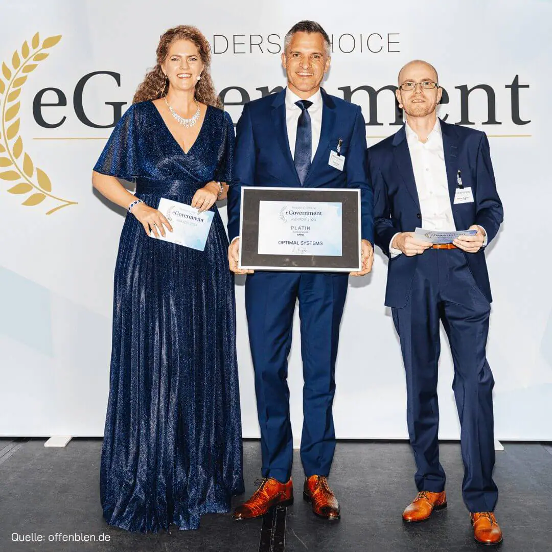 Drei Personen stehen auf einer Bühne und halten ein Zertifikat in den Händen. Die Frau links trägt ein blaues Kleid, die beiden Männer tragen Anzüge. Sie stehen vor einer Kulisse, auf der das Thema „eGovernment“ teilweise zu sehen ist.