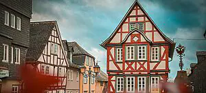 Eine malerische Straßenszene mit mehreren traditionellen Fachwerkhäusern mit rot-weißen Mustern. Der Himmel ist mit ein paar Wolken bedeckt und vor den Gebäuden steht ein Laternenpfahl. Eine verwitterte Statue oder Dekoration ist teilweise sichtbar.