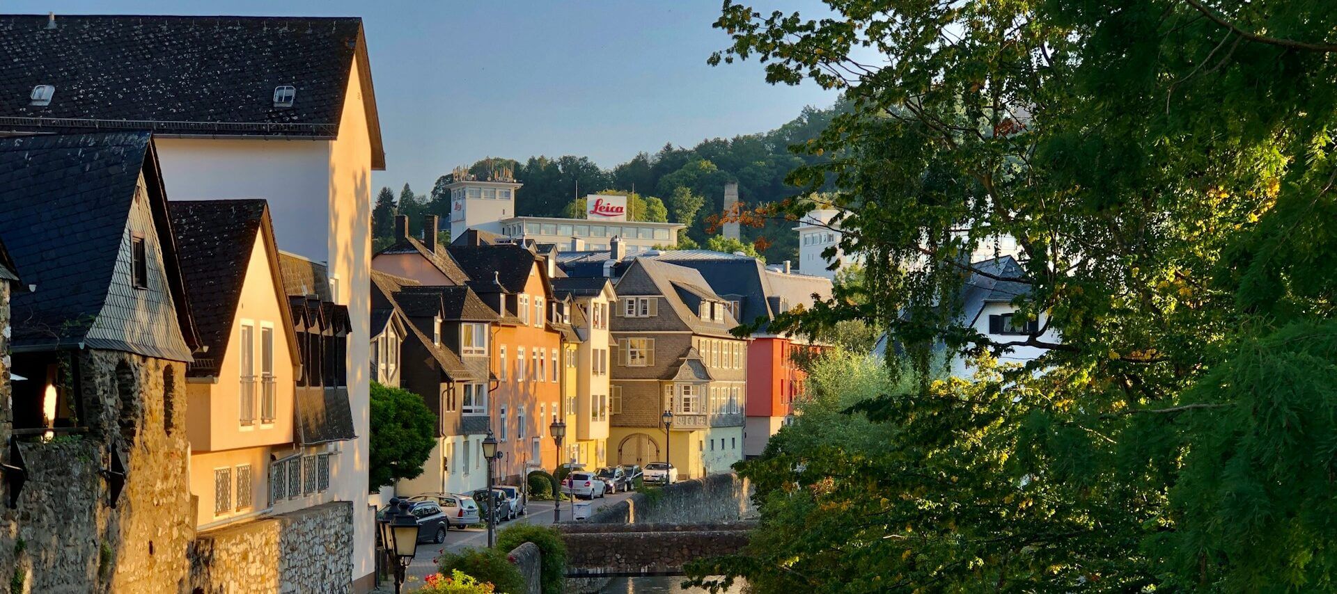 Eine malerische Straßenszene in einer malerischen Stadt mit farbenfrohen Gebäuden entlang einer schmalen Straße. Im Hintergrund ist ein markantes Gebäude mit einem „Leica“-Schild zu sehen. Üppige grüne Bäume rahmen die rechte Seite des Bildes unter einem klaren Himmel ein.