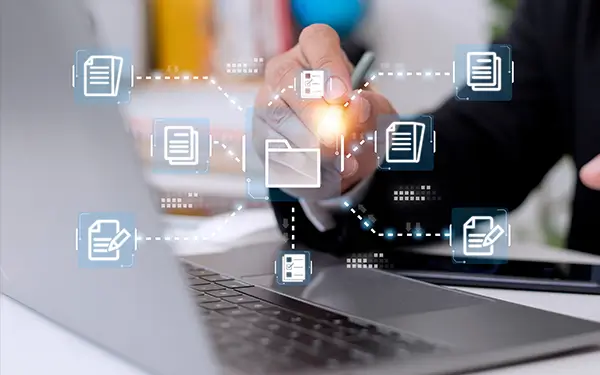 Eine Person hält einen Stift, während sie einen Laptop verwendet. Über dem Bild werden durch gepunktete Linien verbundene Symbole digitaler Dokumente angezeigt, die Dokumentenverwaltung oder den digitalen Arbeitsablauf symbolisieren.