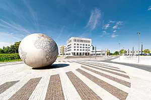 Eine Metallkugel mit strukturierten Mustern steht auf einem Kreisverkehr vor einem modernen weißen Bürogebäude unter einem klaren blauen Himmel.