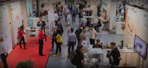 Auf einer belebten Messefläche interagieren Aussteller und Besucher an verschiedenen Ständen. Einige Ausstellungen sind mit Postern und Bildschirmen ausgestattet. Die Leute unterhalten sich und im Hintergrund sind Sitzbereiche und zusätzliche Exponate zu sehen.