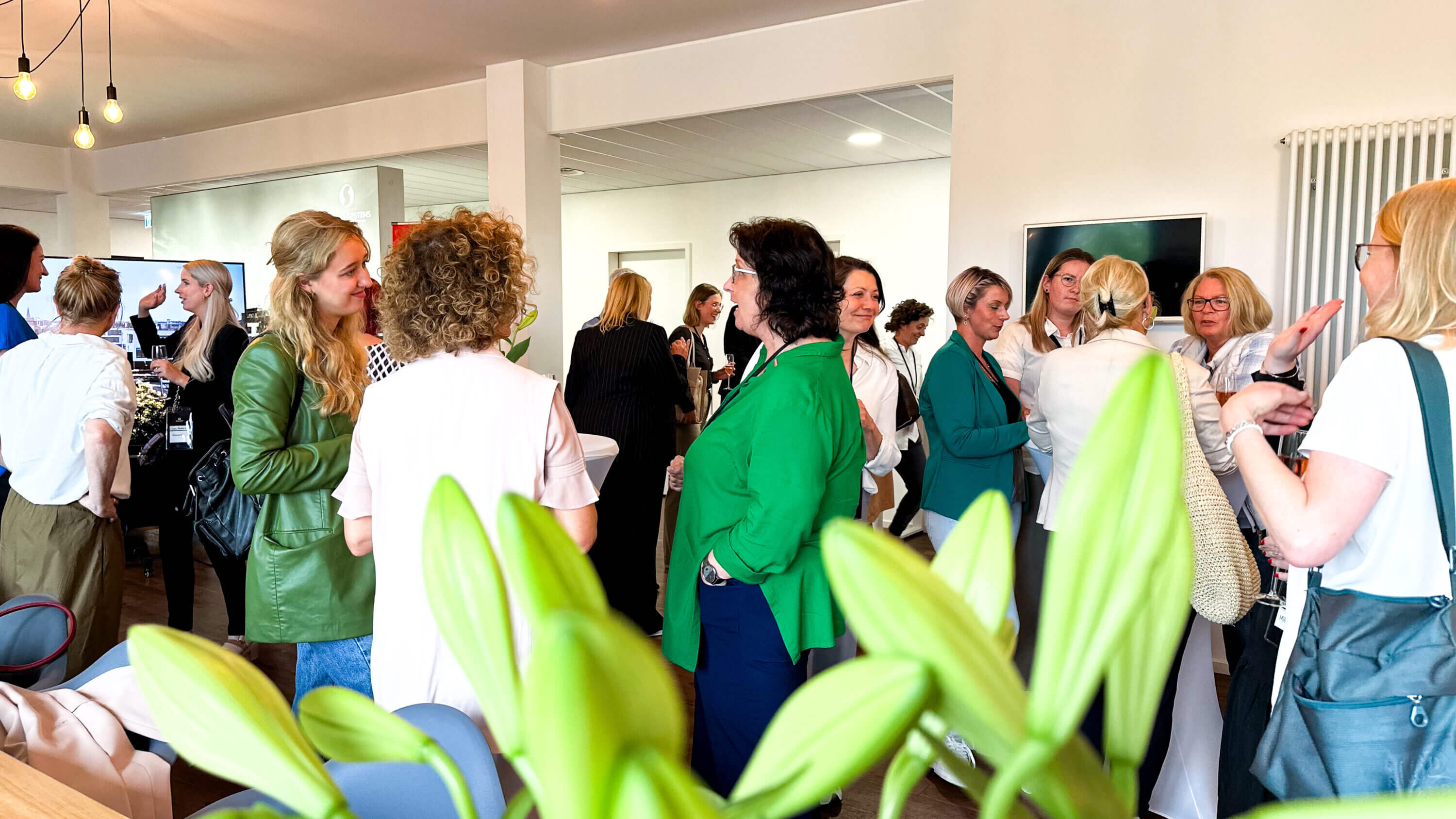 Eine Gruppe von Menschen, hauptsächlich Frauen, unterhält sich in einem hell erleuchteten Raum. Viele tragen Business- oder Freizeitkleidung, einige halten Getränke in der Hand. Im Vordergrund sind grüne Blätter zu sehen, und der Raum ist modern gestaltet und mit Hängelampen ausgestattet.