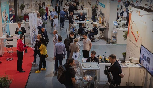 Eine belebte Messehalle mit Ständen und Menschen, die sich unterhalten. Einige Besucher stehen an verschiedenen Ständen, während andere an Tischen sitzen. Bildschirme und Werbematerialien sind sichtbar. Die Kulisse ist eine gut beleuchtete Ausstellungshalle.