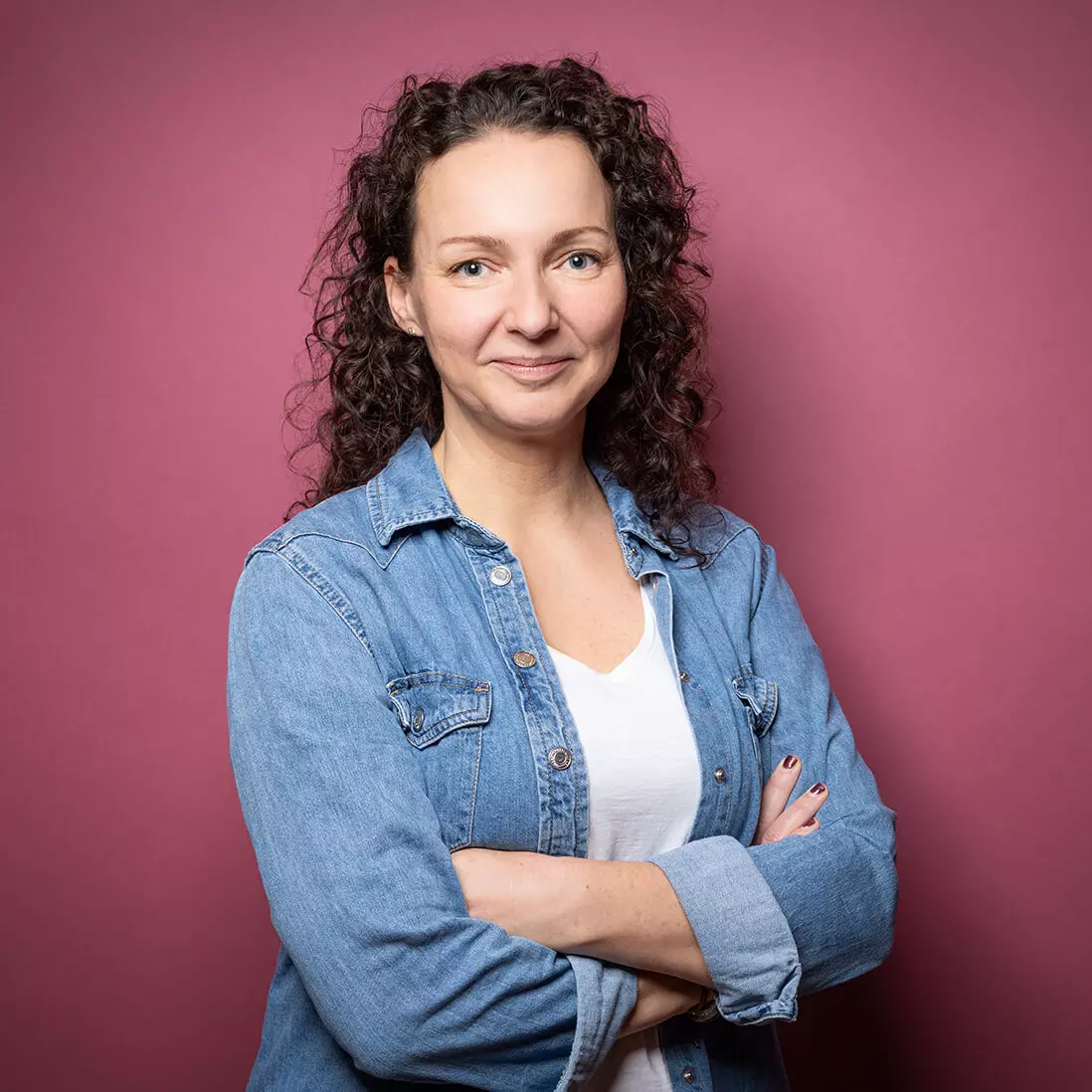 Eine Frau mit lockigem Haar steht vor einem kastanienbraunen Hintergrund und trägt eine Jeansjacke über einem weißen Hemd. Sie lächelt leicht und hat die Arme verschränkt.