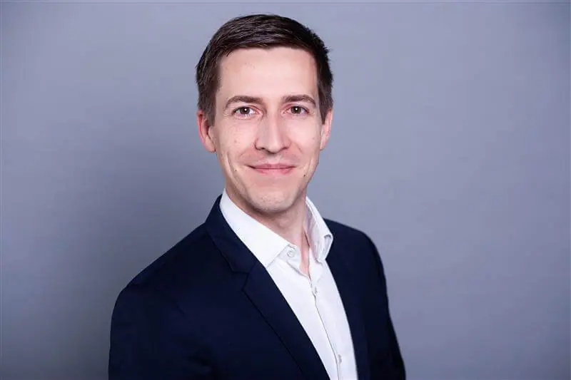Ein Mann mit kurzen braunen Haaren trägt ein dunkles Jackett über einem weißen Hemd. Er lächelt leicht und steht vor einem schlichten grauen Hintergrund.