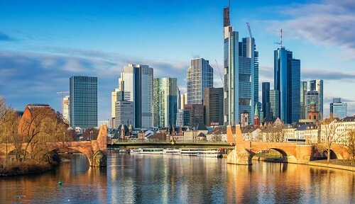 Moderne Wolkenkratzer und historische Gebäude säumen einen Fluss, der von einer Steinbrücke unter einem blauen Himmel mit vereinzelten Wolken überspannt wird.