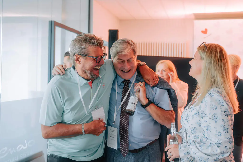 Zwei Männer mit Getränken lachen zusammen, einer hat den Arm um den anderen gelegt. Eine blonde Frau mit Brille steht bei der Indoor-Veranstaltung in der Nähe.