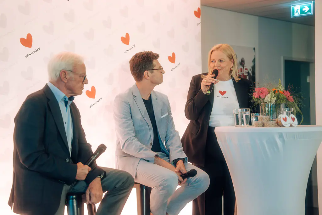 Drei Personen unterhalten sich an einem Tisch mit Mikrofonen, Blumenschmuck, Gläsern und einer weißen Wand mit Herzlogos im Hintergrund.