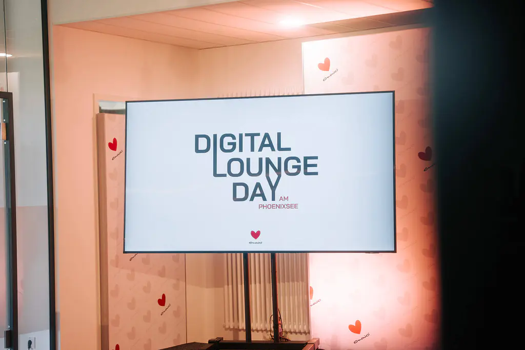 Bildschirm mit "DIGITAL LOUNGE DAY AM PHOENIXSEE" in schwarzer Schrift; helle Wand mit roten Herzsymbolen im Hintergrund.