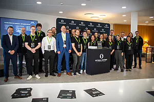 Etwa zwanzig Personen in schwarzen Hemden mit grünen Schlüsselbändern stehen an einem "Optimal Systems"-Stand; auf einem Tisch in der Nähe liegen Materialien.