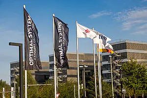 Vier Flaggen - zwei schwarze mit "OPTIMAL SYSTEMS", eine weiße mit Logos und die deutsche Flagge - wehen vor einem modernen Bürogebäude.