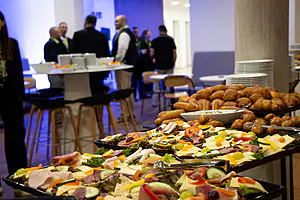 Vorne steht ein Buffet mit verschiedenen Wurst-, Käse- und Brotsorten; die Leute unterhalten sich an Stehtischen und Tellern in einer modernen Inneneinrichtung.