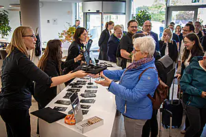 Eine Frau in einer blauen Jacke erhält bei der Anmeldung eine Tragetasche, während die anderen warten und Namensschilder und Broschüren auf dem Tresen liegen.