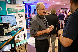 Ein Mann in einem grauen Hemd unterhält sich mit zwei Personen in der Nähe eines Kodak Alaris Standes bei einer belebten Veranstaltung in einer Halle. Andere sind im Hintergrund zu sehen.