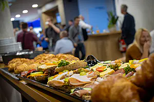 Eine Platte mit Sandwiches und Gebäck steht auf einem Tisch, während sich die Leute in einem modernen Innenraum im Hintergrund tummeln.