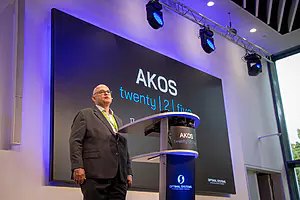 Ein Mann im Anzug präsentiert an einem Podium; hinter ihm zeigt ein Bildschirm "AKOS twenty | 2 | five" und das Logo von Optimal Systems in einem modernen Raum.