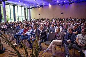 Das aufmerksame Publikum sitzt in einem modernen, gut beleuchteten Konferenzraum mit Holzwänden, großen Fenstern und violetter Deckenbeleuchtung.