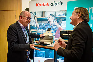 Zwei Männer in Geschäftskleidung plaudern und lächeln an einem Stand von Kodak Alaris, einer deutet auf eine Büroausstattung mit Werbematerialien dahinter.