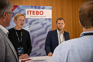 Zwei Männer an einem Stand der ITEBO Unternehmensgruppe unterhalten sich mit zwei Besuchern; Broschüren und Ausweise sind sichtbar.