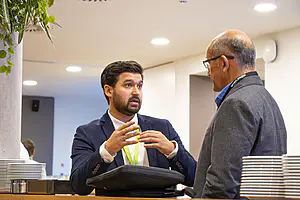 Zwei Männer in Geschäftskleidung und mit Konferenzschildern unterhalten sich in einem modernen Innenraum neben einem Tresen mit gestapelten Tellern.