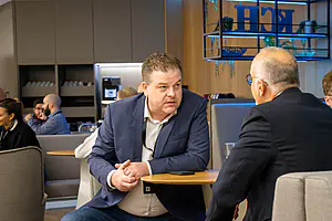 Zwei Männer in Geschäftsanzügen unterhalten sich ernsthaft an einem kleinen Tisch in einem hellen Café, während sich andere im Hintergrund unterhalten.