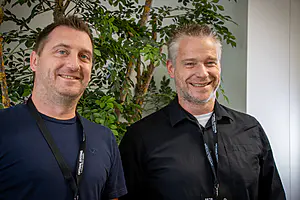 Zwei lächelnde Männer in legeren Hemden und schwarzen Schlüsselbändern stehen zusammen vor einer begrünten Zimmerpflanze und einer weißen Wand.