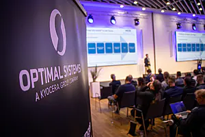 Banner mit dem Logo von "Optimal Systems, a Kyocera Group Company"; Personen beobachten einen Moderator und eine Folie in einem Konferenzraum.