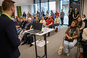 Der Redner auf dem Podium mit Laptop spricht zu den sitzenden Zuhörern; einige stehen im hinteren Bereich. Große Fenster erfüllen den Konferenzraum mit Licht.