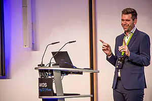 Ein lächelnder Mann im Anzug steht an einem Podium mit Mikrofonen und einem Laptop und gestikuliert mit beiden Händen. Glatte Wand hinter ihm.