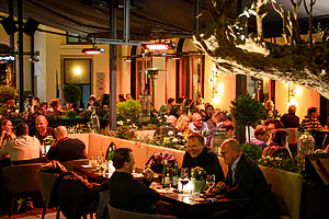 Nachts ist das Restaurant unter freiem Himmel, mit warmen Lichtern und Heizkörpern, umgeben von Pflanzen in einer lebhaften, entspannten Atmosphäre.