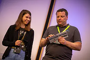 Eine Frau mit Mikrofon steht neben einem Mann mit Headset, der eine Broschüre hält; beide schauen bei einer Veranstaltung nach vorne.