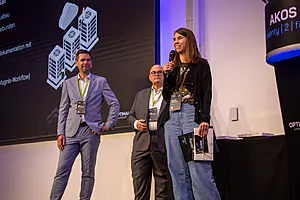 Drei Moderatoren auf der Bühne - eine Frau mit einem Mikrofon, zwei Männer in der Nähe; großer Bildschirm mit Diagrammen und Text im Hintergrund.