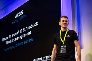 Mann in schwarzem Poloshirt und Schlüsselband steht neben einem Bildschirm, auf dem die Präsentation "Neues in enaio® 12" von Florian Strüber, OPTIMAL SYSTEMS, läuft.