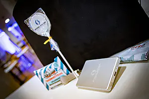 Ein silbernes Ladegerät, Bonbons, Minzbonbons, ein USB-Stick und eine Broschüre liegen auf einem weißen Tisch vor einer schwarzen Tasche.