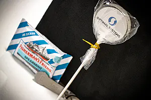 Fisherman's Friend-Minze, ein weißer "OPTIMAL SYSTEMS"-Lutscher und ein grauer Plastikartikel liegen auf einer dunklen Fläche.