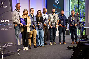 Acht Preisträger mit Plaketten stehen auf der Bühne vor einem Bildschirm mit der Aufschrift "AKOS twenty | 2 | five", rechts daneben ein Podium.