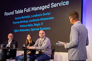 Vier Personen auf der Bühne in einer Diskussionsrunde; drei sitzen an einem Tisch, eine steht. Die Folie dahinter zeigt Namen und Organisationen.