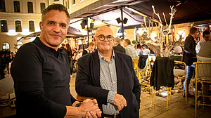 Zwei Männer, einer im Polohemd, der andere in einer Anzugsjacke, in einem belebten, warm beleuchteten modernen Restaurant oder einer Bar.