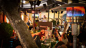 Ein DJ spielt an Ressos Stand in einem belebten Außenrestaurant, umgeben von Gästen, Pflanzen und Heizkörpern.