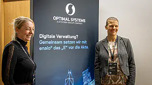 Zwei Frauen in Geschäftskleidung lächeln in einem Büro neben einem "OPTIMAL SYSTEMS"-Display mit deutschem Text.