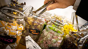 Eine Hand wählt mit einer Zange Süßigkeiten aus einem Glas mit der Aufschrift "Kein Schwein" aus, inmitten von Gläsern mit verschiedenen verpackten und unverpackten Süßigkeiten.