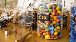 Auf der Holztheke stehen Gläser mit verschiedenen Bonbons und Schokoladennüssen, die mit Kreideschildern beschriftet sind; daneben liegt eine Metallzange.